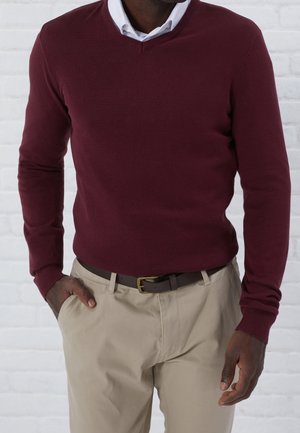 Burgunderfarbener V-Ausschnitt-Pullover aus Strickstoff, mit gerippten Bündchen und Saum. Kombiniert mit hellbeigen Hosen und einem braunen Gürtel.