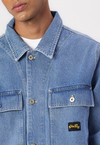 Blue denim jacket featuring a standard collar, two front pockets with flaps, yellow stitching accents, and a black label on the left pocket.