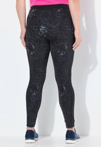 Kvinna iklädd svarta leggings med blommönster och blå sneakers, stående vänd mot en vit vägg, med händerna vilande på höfterna.