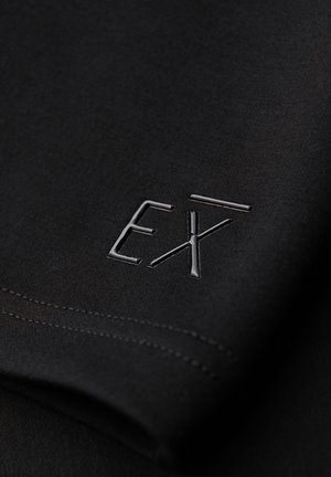 Tissu noir avec logo "EXT" brillant en relief près du bord cousu, montrant la texture et les détails du vêtement en gros plan.