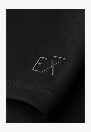 Tissu noir avec logo "EXT" brillant en relief près du bord cousu, montrant la texture et les détails du vêtement en gros plan.