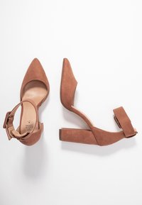 Brown suede high heels with a pointed toe, adjustable ankle strap, and block heel. Smooth texture and minimalistic design.