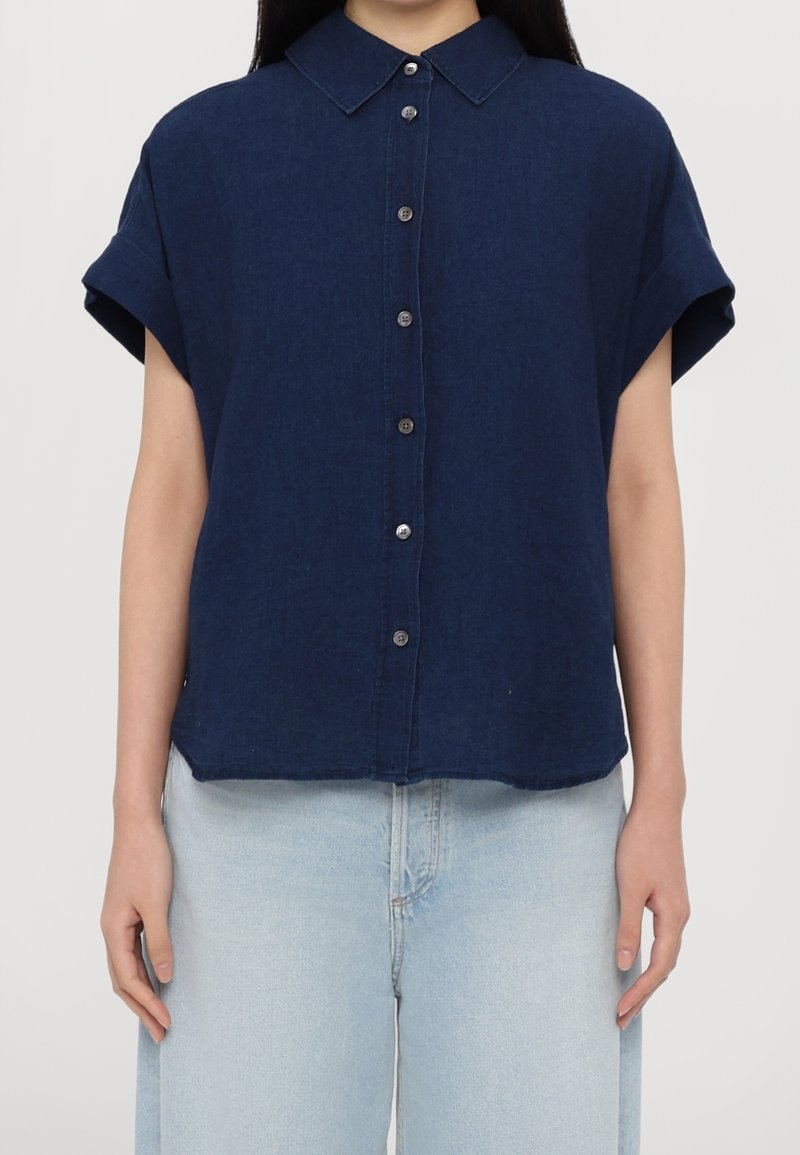 Personne portant une chemise bleu foncé à manches courtes avec col et boutons, associée à un jean taille haute bleu clair sur un fond uni.