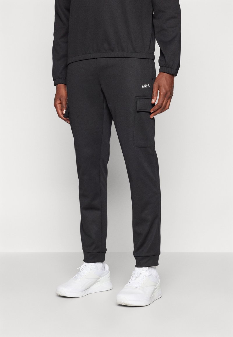 Homme portant un pantalon jogger cargo noir et des baskets blanches, debout sur un fond uni avec les mains détendues le long du corps.