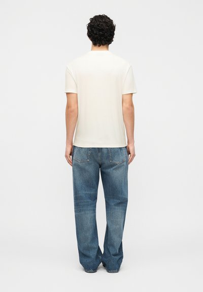 Άνδρας με κυματιστά μαλλιά στέκεται με την πλάτη γυρισμένη φορώντας απλό λευκό κοντομάνικο πουκάμισο και φαρδιά μπλε τζιν παντελόνια.