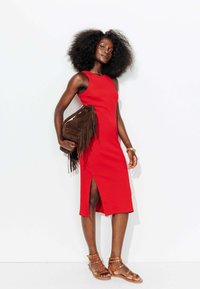 Robe rouge ajustée avec une fente sur le côté, portée avec un sac à franges marron et des sandales cloutées marron clair. Tissu lisse, design minimaliste.