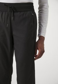 Mustat venyvät housut, joissa on uritettu vyötärö, kiristysnauha ja sivuvetoketjulliset taskut. Sileä pinta ja slim-fit-malli.