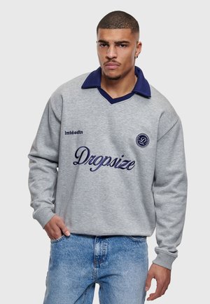 Młody mężczyzna z krótkimi włosami, ubrany w szarą bluzę z granatowym kołnierzem i napisem "Dropsize" na przodzie, w połączeniu z jasnoniebieskimi dżinsami.