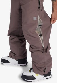 Pantalons de neige imperméables marron avec des aérations en maille zippées et le logo DC, accompagnés de chaussures de neige noir et blanc avec une semelle épaisse.