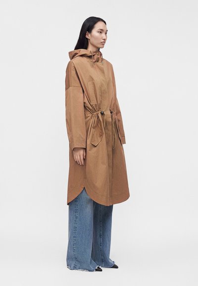 Femme portant un long manteau marron à capuche avec des cordons et un jean bleu à jambes larges, debout devant un fond clair uni.