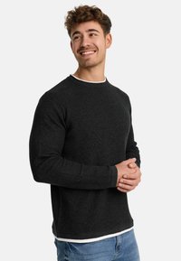 Suéter negro texturizado con cuello redondo, mangas largas y un sutil acento blanco en el collar, superpuesto a una camisa blanca, combinado con jeans azules.
