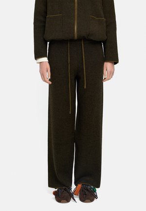 Ciemno-oliwkowe spodnie o szerokich nogawkach z sznurkiem w talii, noszone z pasującą kurtką na zamek i brązowymi trampkami ozdobionymi kolorowymi frędzlami.