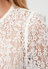 Λευκό πουκάμισο δαντέλας με λουλουδάτο μοτίβο, διάφανο ύφασμα και περίπλοκες λεπτομέρειες. Διαθέτει λαιμόκοψη υψηλού τύπου και κοντά μανίκια.