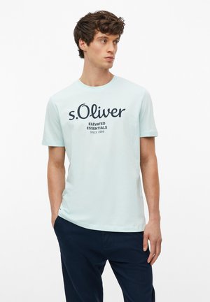Młody mężczyzna noszący jasnoniebieską koszulkę s.Oliver z napisem "Elevated Essentials Since 1969" i ciemnoniebieskie spodnie, patrzy na bok.