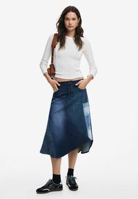 Jeansrock mit asymmetrischem Schnitt, verschiedenen Blautönen, vorderen Knöpfen und einer aufgesetzten Tasche. Kombiniert mit einem figurbetonten weißen Oberteil und Sneakers.