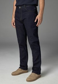Dunkelblaue Denim-Jeans mit geradem Schnitt, sichtbaren Nähten, Fronttaschen und Knopfverschluss. Dazu werden hellbraune Sneakers getragen.