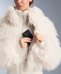 Jasno beżowa kurtka z puszystymi, teksturowanymi rękawami z futra, z wysokim kołnierzem i przednim zamkiem; do środka wkładany jest smartfon.