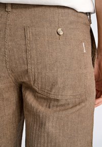 Pantalones marrones con patrón de espiga, con un bolsillo trasero y un cierre de botón, hechos de una tela texturizada. Presentan una pequeña etiqueta de la marca.