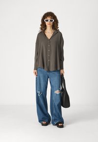 Camisa oversized cinza escura com botões, jeans blue distressed de perna larga, mala preta e óculos de sol brancos. Tecido suave, ajuste relaxado, grande aba.