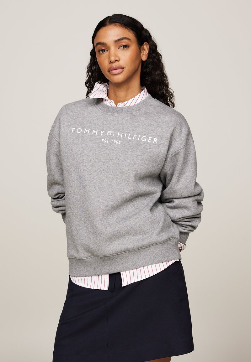 Felpa grigia con polsini e orlo a coste, decorata con la scritta bianca "TOMMY HILFIGER EST. 1985". Indossata sopra una camicia a righe con colletto e una gonna blu navy.