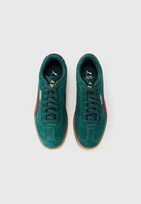 O pereche de pantofi sport Puma din suede verde teal cu dungi burgundy, logo-uri aurii și talpă din cauciuc de culoare bej, văzuți dintr-o perspectivă de sus pe un fundal alb.