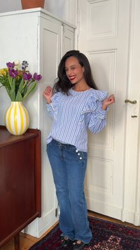 Γυναίκα που χαμογελά με μακριά σκούρα μαλλιά, φοράει μπλε και λευκή ριγέ μπλούζα με βολάν και μπλε τζιν, στέκεται κοντά σε ένα ντουλάπι με βάζο με κίτρινες ρίγες.