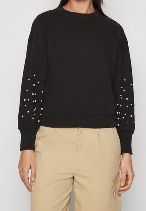 Mujer con top negro de manga larga con adornos de perlas en las mangas y pantalones beige con bolsillos laterales, de pie frente a un fondo liso.