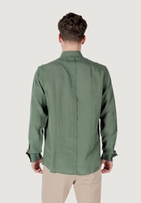 Camicia verde a maniche lunghe in tessuto liscio con colletto alla cinese, caratterizzata da una cucitura centrale e polsini con bottoni. Indossata con pantaloni beige.