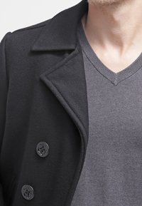 Czarny wełniany płaszcz z wyciętym kołnierzem, zapinany na trzy przednie guziki, noszony na szarym swetrze z dekoltem w serek. Gładka faktura i dopasowany krój.