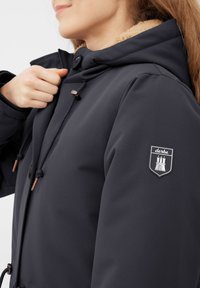 Žena upravuje zip tmavé mikiny s kapucí, které je podšité béžovou látkou a má nášivku "derbe" na horním rukávu.