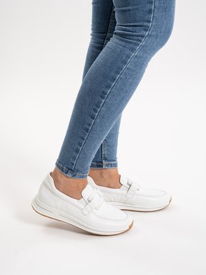 Person trägt weiße Slip-On-Sneaker und blaue Skinny-Jeans, steht vor weißem Hintergrund.
