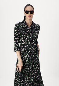 Robe noire avec un motif de feuilles en rose et vert, à manches longues, fermeture à boutons sur le devant, et coupe fluide. Portée avec des lunettes de soleil noires en forme de chat.