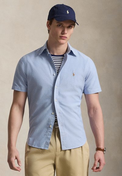 Jaunas vyras dėvi tamsiai mėlyną beisbolo kepurę, šviesiai mėlyną trumpomis rankovėmis marškinius su sagomis ant tamsiai mėlynos dryžuotos marškinėlių, smėlio spalvos kelnes ir riešo laikrodį.