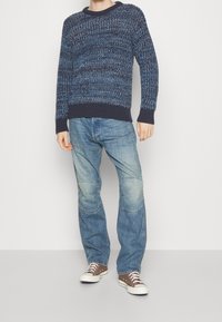 Objemný pletený svetr v modrých tónech, kulatý výstřih, žebrované manžety, v kombinaci se světle modrými džíny s kolenními záplaty a hnědými teniskami.