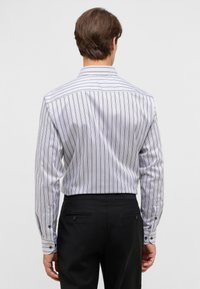Man die met zijn rug naar de camera staat, gekleed in een wit shirt met dunne verticale zwarte strepen en zwarte pantalon, tegen een effen lichte achtergrond.
