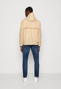Beige letvægtsjakke med hætte, rygventilation og elastiske manchetter; parret med mørkeblå jeans og hvide sneakers.