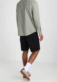 Osoba ubrana w jasnoszary, długi rękaw, czarne szorty, białe skarpetki i jasnoszare buty sportowe, stojąca tyłem do kamery.