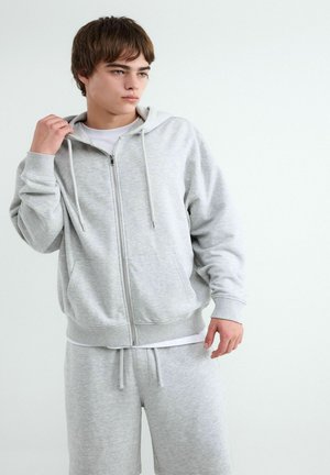 Jonge man die de kraag van een lichtgrijze hoodie met rits aanpast, met bijpassende joggingbroek en een wit shirt eronder.