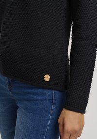 Zwarte gestructureerde gebreide trui met een ronde houten logo-accent aan de zoom, gecombineerd met blauwe denim jeans.