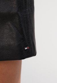 Svart läderkjol med en texturerad yta. Har en liten metall-logotag med röda, vita och blå accenter nära fållen.