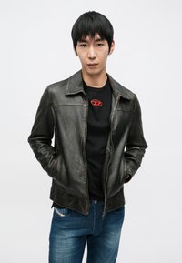 Giacca nera in pelle con una texture liscia, abbinata a una camicia con un logo rosso e jeans blu, su uno sfondo bianco uniforme.