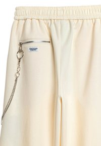Pantalones de chándal color crema con cintura elástica, que cuentan con un bolsillo con cremallera y un acento de cadena de metal. Material suave y ajuste holgado.