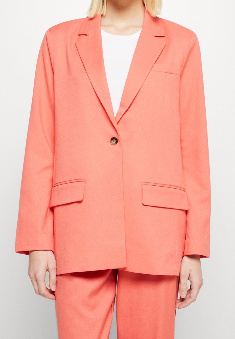 Personne portant un blazer corail rose ajusté avec des revers crantés, une fermeture à un bouton, des poches à rabat et un pantalon assorti sur un top blanc.