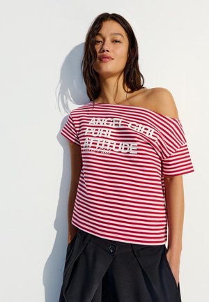 Frau mit langem dunklem Haar, die ein rot-weiß gestreiftes, schulterfreies Oberteil mit Text und schwarze Hosen trägt, steht vor einer hellen Wand.