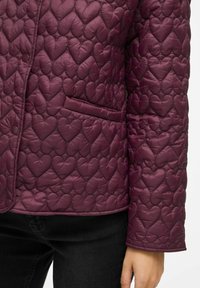 Gequilted jasje in diep bordeauxrood met hartvormige patronen, voorzien van een enkele zak en knoopsluiting, gemaakt van glanzend materiaal.