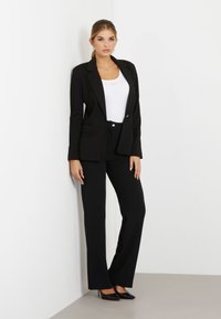 Blazer nero su misura sopra una blusa bianca, abbinato a pantaloni neri. Presenta linee eleganti, rever classici e tacchi alti per un look raffinato.