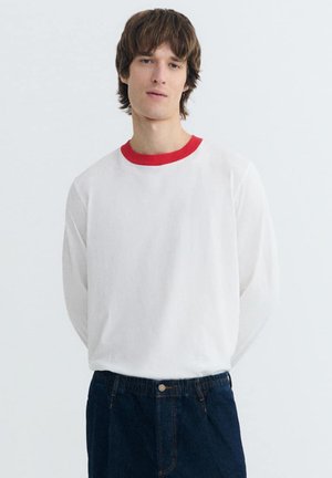 Junger Mann mit braunen Haaren, der ein weißes Langarmshirt mit rotem Kragen und dunkelblaue Jeans trägt, vor einem schlichten hellen Hintergrund stehend.