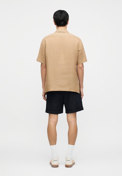 Άνδρας που στέκεται με την πλάτη στραμμένη, φοράει μπεζ κοντομάνικο πουκάμισο, μαύρα σορτς, λευκές κάλτσες και μπεζ αθλητικά παπούτσια μπροστά σε απλό φόντο.