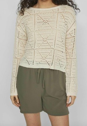 Femme portant un pull en crochet à manches longues de couleur crème associé à un short kaki à cordon de serrage, sur un fond uni.