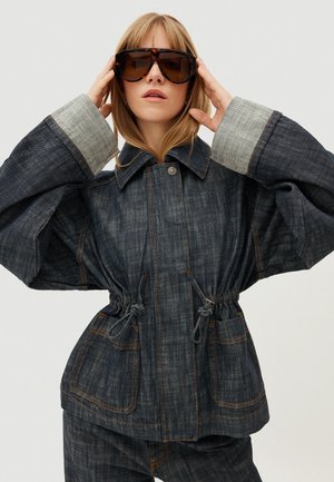 Jonge vrouw die een oversized zonnebril verstelt en een donker denimjack draagt met grote manchetten en een tunnelkoord in de taille.